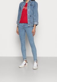 Denimjack, lichtblauw, met twee voorzakken en knoopsluiting. Rode sweatshirt eronder, slim-fit lichtblauwe jeans, witte sneakers met accenten.