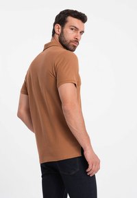 Hnědý polo tričko z měkké tkaniny s klasickým límečkem, krátkými rukávy a přiléhavým střihem. Nosené s tmavými džínami.