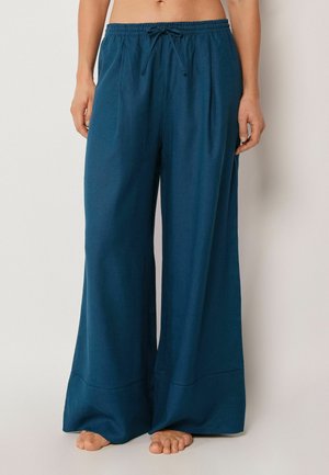 Blå bukser med vide ben og elastisk taljebånd med snører, båret barfodet, vist fra talje til fødder mod en ensfarvet baggrund.