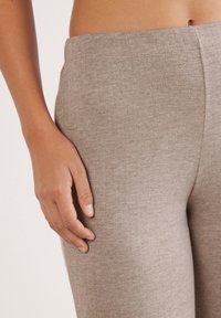 Gros plan sur la main d'une personne reposant sur un pantalon beige texturé, montrant le détail du tissu et une partie du torse avec un nombril visible.