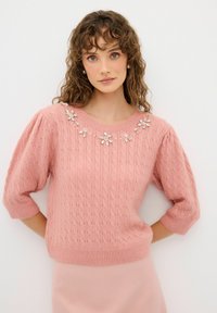 Pull en tricot rose avec un ourlet côtelé et des manches bouffantes courtes. Décoré de garnitures florales ornementales au niveau de l'encolure.