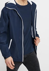 Laivastonsininen tuulipuku takki fleece-vuorattu huppu, valkoinen vetskari ja etutaskut, yhdistettynä laivastonsiniseen T-paitaan ja tummaan farkkuihin.