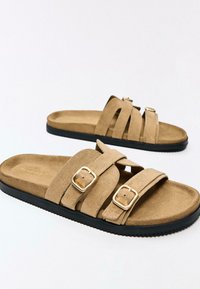 Světle hnědé semišové nazouváky se dvěma nastavitelnými pásky a zlatými přezkami, s texturovanou černou gumovou podrážkou. Měkká stélka pro pohodlí.