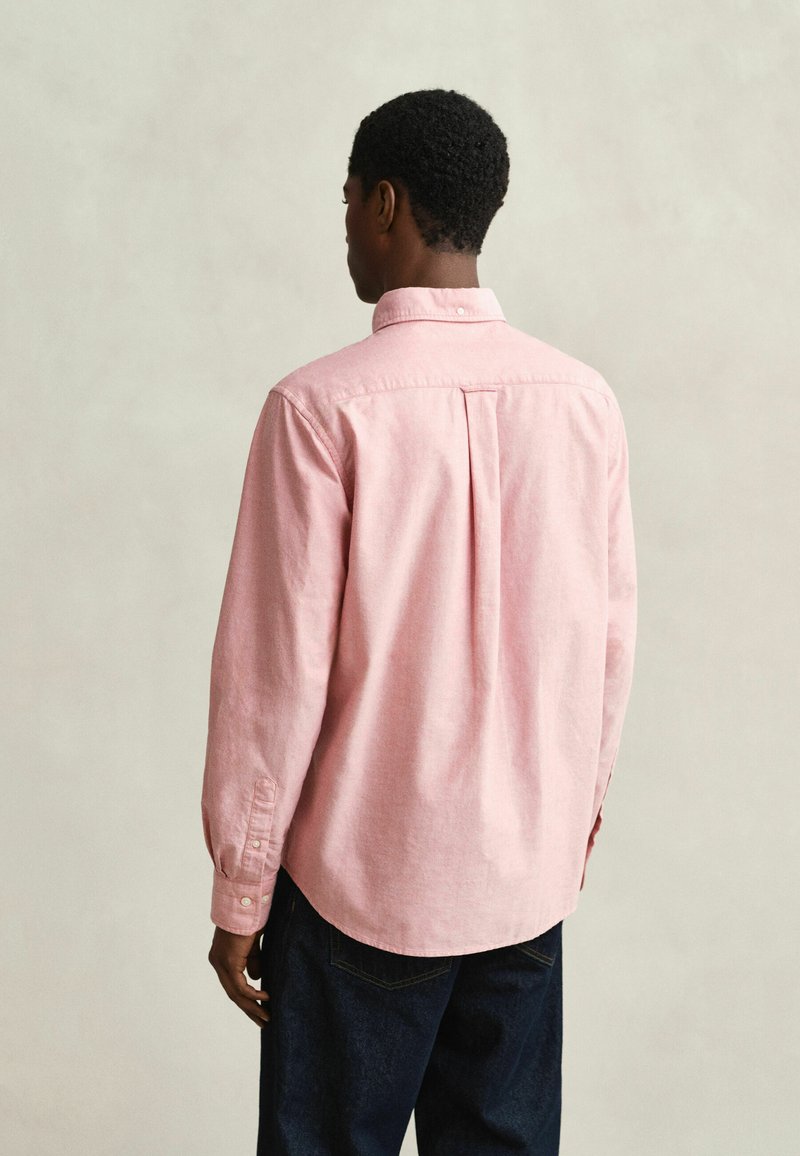 Hombre visto desde atrás con una camisa de manga larga color rosa claro abotonada y vaqueros azul oscuro sobre un fondo claro y liso.