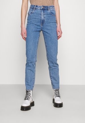 Personne portant un jean taille haute bleu clair et des bottes de combat blanches à lacets, debout devant un fond blanc uni.