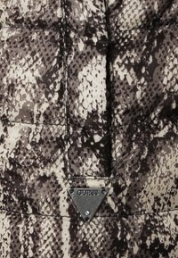 Tissu matelassé imprimé serpent marron et beige avec un logo triangulaire métallique Guess fixé près du centre en bas.