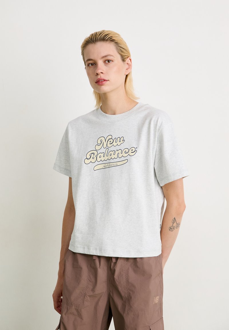 T-shirt en coton gris clair à manches courtes, arborant le logo "New Balance" en écriture marron sur la poitrine ; encolure arrondie, coupe décontractée.