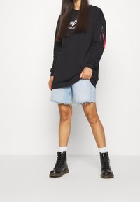 Sweat-shirt noir avec logo, manches longues et poche latérale zippée. Short en jean bleu clair avec ourlet effiloché. Bottes noires à lacets et chaussettes blanches.