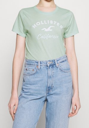 Osoba nosząca jasnorozmową koszulkę Hollister California włożoną do jasnoniebieskich spodni z wysokim stanem na jednolitym tle.