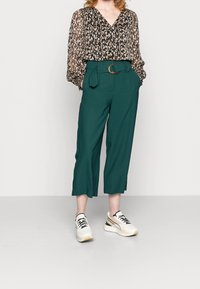 Blusa nera trasparente con motivo floreale e scollatura a V. Pantaloni cropped verdi con una fibbia di cintura in legno. Sneakers bianche con accenti neri.