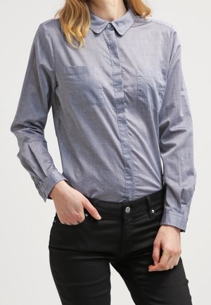 Nainen, jolla on vaaleansininen napillinen pitkähiha-paita, jossa on kaksi rinnantaskua, ja mustat housut, käsi taskun lähellä leväten.