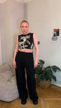 Mustast lõigatud tanktop graafilise disainiga, kombineeritud lahtiste mustade pükstega ja massiivsete mustade kingadega. Taustal punutud ümmargune taimealus.