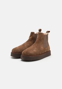 Botas Chelsea de gamuza marrón con paneles laterales elásticos, punta redondeada y una suela de goma marrón gruesa y texturizada. Incluye un tirador trasero.