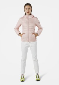 Giacca puffer rosa chiaro con cappuccio, zip frontale, texture trapuntata e tasche laterali. Indossata con pantaloni bianchi e sneakers con accenti verdi.