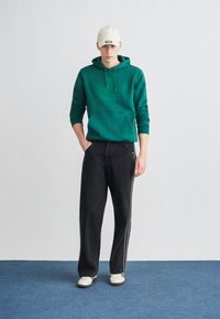 Πράσινο φούτερ με κουκούλα και μπροστινή τσέπη, μαύρο φαρδύ τζιν με ραφές στο πλάι και λευκά αθλητικά παπούτσια. Ένα ουδέτερου χρώματος καπέλο ολοκληρώνει το σύνολο.