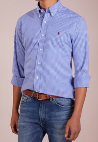 Camicia button-down a quadri blu e bianchi con colletto, maniche arrotolate e un piccolo logo ricamato. Abbinata a jeans in denim e una cintura marrone.