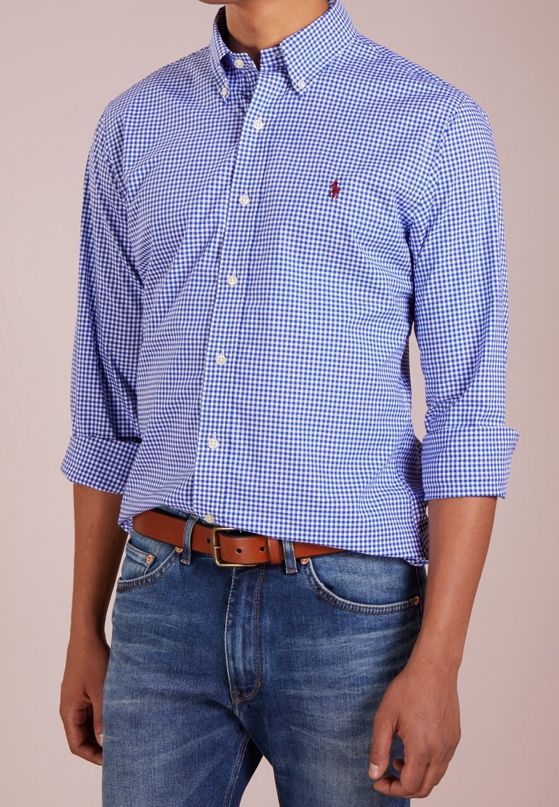 Camicia button-down a quadri blu e bianchi con colletto, maniche arrotolate e un piccolo logo ricamato. Abbinata a jeans in denim e una cintura marrone.