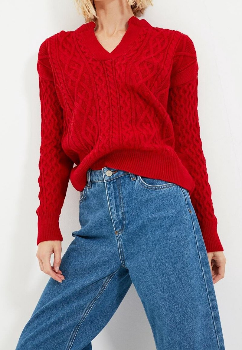 Personne portant un pull rouge à col en V en maille torsadée et un jean bleu ample taille haute sur un fond clair uni.