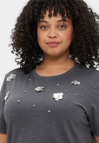 Grå T-skjorte laget av mykt stoff med sølvfargede blomsterdekorasjoner og klare rhinestones. Rund hals og korte ermer.