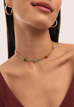 Mujer con pendientes de aro dorados y un collar de cadena dorada con piedras ovaladas de colores, sobre hombros desnudos y una blusa granate.
