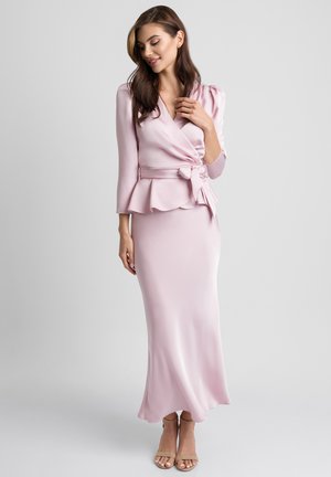 Kobieta w jasnoróżowej satynowej bluzce zapinanej na zakładkę z kokardą i dopasowanej długiej spódnicy, stojąca z jedną ręką dotykającą kołnierzyka bluzki.