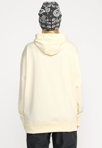 Hoodie crème avec une coupe décontractée, présentant une grande capuche et un ourlet côtelé. Associé à un bonnet noir à motifs.