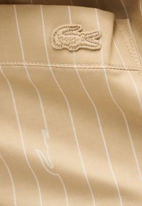 Tissu beige avec des fines rayures verticales blanches et un logo crocodile beige cousu près d'une couture.