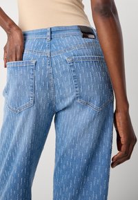 Person som har på seg lyseblå teksturerte jeans med baklommer, en hånd plassert i en lomme, og en beige topp som er stukket inn.