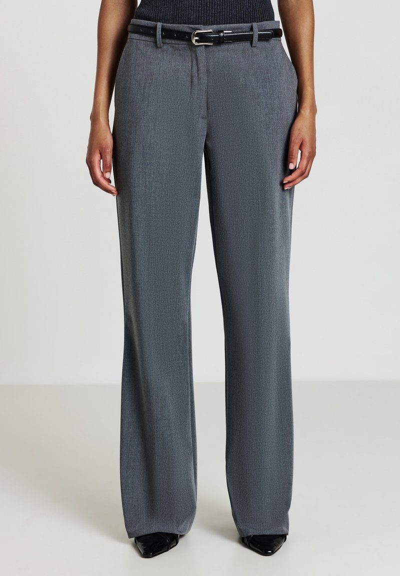 Femme portant un pantalon gris taille haute à jambes larges, ceinture noire et chaussures noires à bouts pointus, debout devant un fond uni.