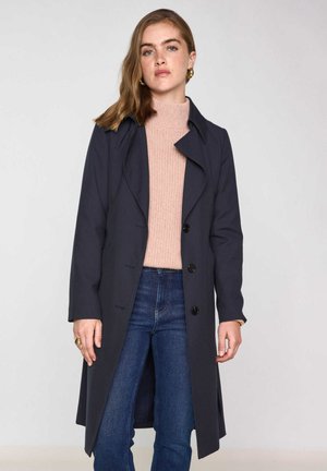 Trench coat navy con colletto, design doppiopetto, bottoni neri e maniche lunghe, indossato sopra un maglione a coste rosa chiaro e jeans.