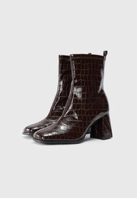 Botas de tobillo de cuero sintético marrón con estampado de cocodrilo, punta cuadrada, tacón alto y borde superior cosido. Cuenta con un tirador en la parte trasera para facilitar el uso.