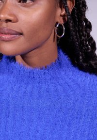 Un pull bleu duveteux avec un col montant et une texture côtelée. Présente un gros plan d'une boucle d'oreille en argent contre des cheveux tressés.
