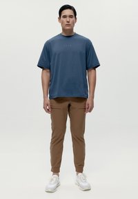 Šedoblá lé bavlněné tričko s krátkým rukávem a kulatým výstřihem. Hnědé tapered jogger kalhoty s dvěma předními kapsami a kotníkovými manžetami. Bílé tenisky.