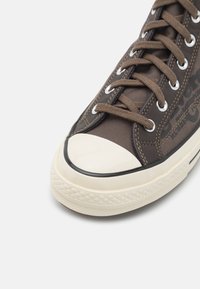 Brązowe płócienne trampki z białą gumową czubką i fakturowaną podeszwą, z brązowymi sznurowadłami, srebrnymi oczkami i subtelnymi detalami po bokach.