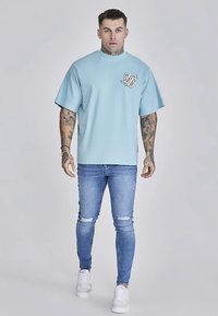 Light blue oversized t-shirt with short sleeves and a front logo, paired with distressed blue jeans and white sneakers.