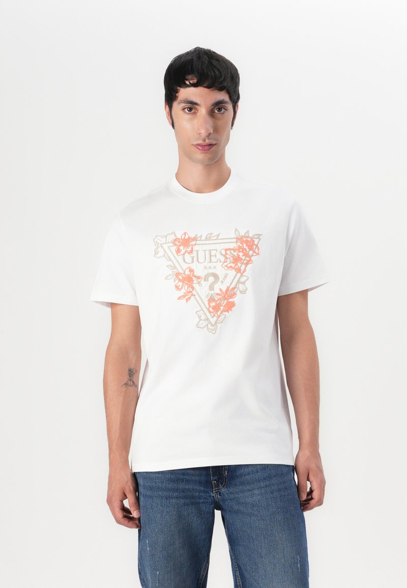 Bílý bavlněný tričko s centrálním grafickým motivem květinových vzorů a textem "GUESS" v světle šedé a oranžové. Klasický design s kulatým výstřihem.