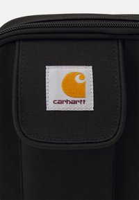 Svart stoffpose med glidelås, med en hvit firkantet lapp som viser en gul logo og teksten "carhartt" i mørkerød.