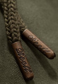 Cuerdas de tiro trenzadas marrones con extremos metálicos grabados con "Reell", descansando sobre un fondo de tejido texturizado verde oliva.