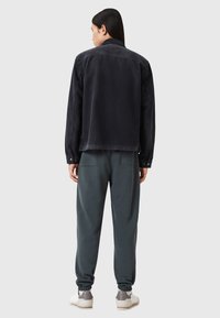 Zwarte fluwelen jas met een gestructureerde kraag, gecombineerd met relaxte grijze sweatpants. Schoeisel heeft grijze en witte accenten.