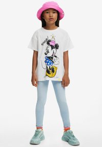 Hvid t-shirt med grafik af en tegneseriefigur, blå prikket nederdel, kombineret med lyse denim leggings og teal sneakers, pink bøttehat.