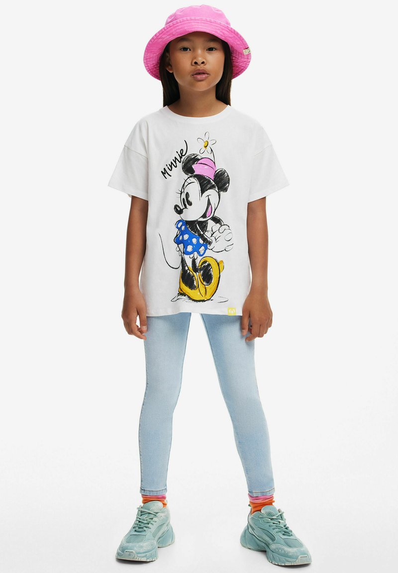 Hvid t-shirt med grafik af en tegneseriefigur, blå prikket nederdel, kombineret med lyse denim leggings og teal sneakers, pink bøttehat.