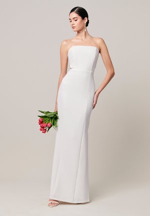 Žena v bielych šatách bez ramienok až po zem, držiaca kyticu ružových tulipánov, stojaca pred jednofarebným béžovým pozadím.