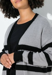 Harmaa ja musta raidallinen neuletakki, rentoa istuvuutta, pitkät hihat, avoin etuosa ja ribbireunat. Pehmeä neulekuitu tarjoaa mukavuutta.