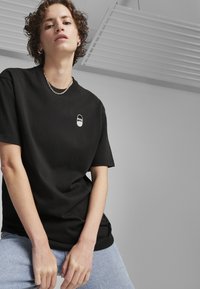 Juodas medvilninis marškinėlis su apvalia iškirpte, ant krūtinės turi mažą baltą Puma logotipą. Modelis dėvi šviesiai mėlynus džinsus.