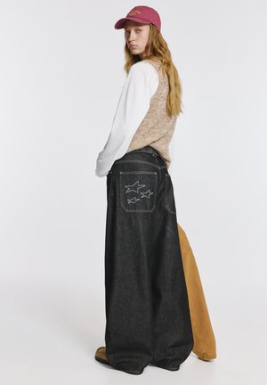 Vide benjeans i mørkt denim med stjernebroderi på baglommerne, kombineret med en beige strikvest og hvid langærmet top. Bourgognefarvet kasket.