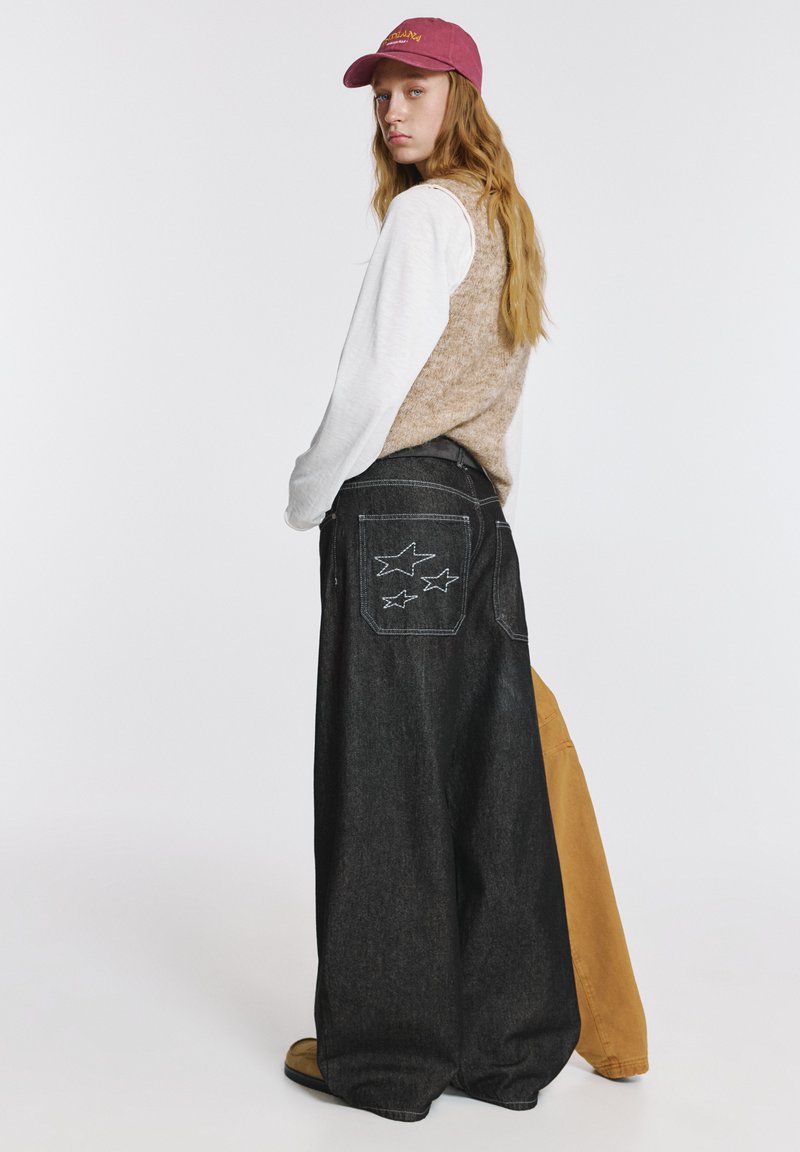 Široke jeans hlače iz temnega džinsa z zvezdastim vezenjem na zadnjih žepih, kombinirane s bež pleteno jopico in belo majico z dolgimi rokavi. Bordo kapa.