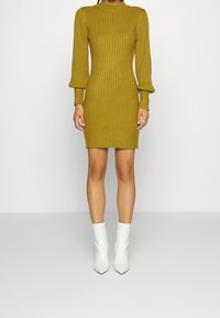 Robe tricotée côtelée moutarde avec de longues manches bouffantes et un col montant, assortie de bottes blanches brillantes à la cheville.