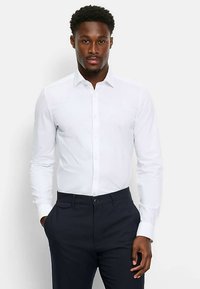 Weißes Langarmhemd mit Button-Down-Kragen, schmal geschnitten, aus glattem Baumwollstoff, kombiniert mit dunkler Hose.