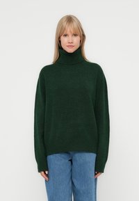 Pull à col roulé vert foncé en matériau tricot doux, avec une coupe ample et des manches longues, associé à un jean taille haute bleu clair.