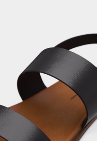 Κοντινό πλάνο μαύρου δερμάτινου σανδαλιού με δύο φαρδιές λουριά πάνω σε καφέ πάτο, σε λευκό φόντο.